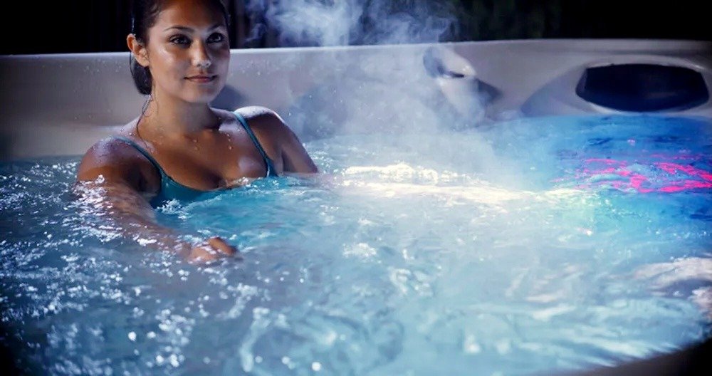 A woman relaxes in a steaming outdoor hot tub with blue LED lighting at night.