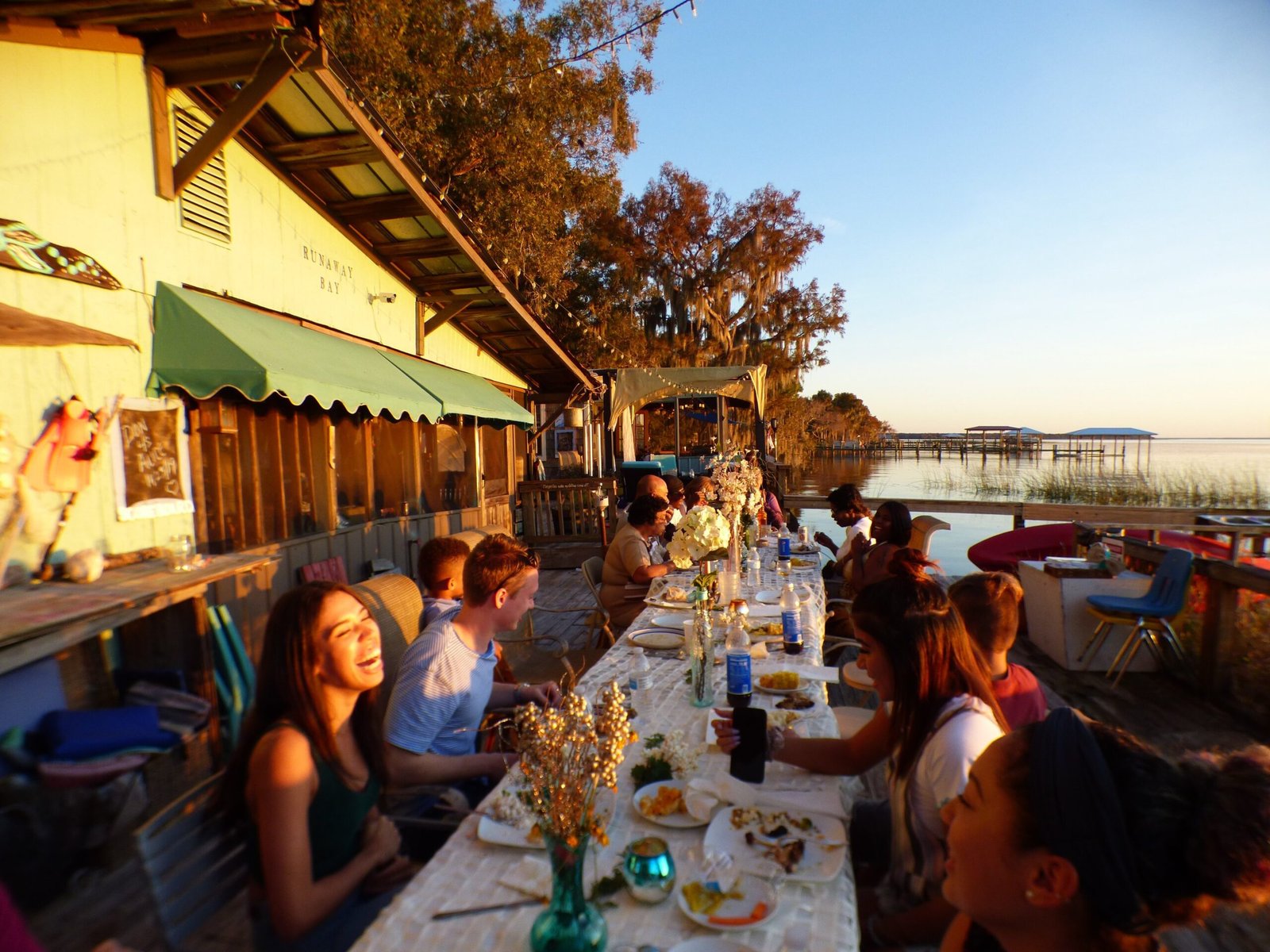 A group of friends enjoying a waterfront dinner party at sunset on a rustic lakeside patio.