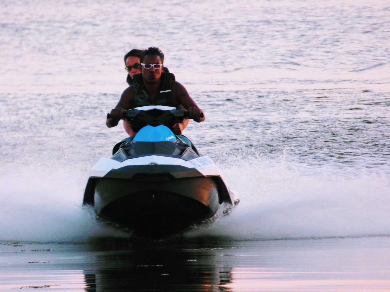 Two people wearing sunglasses riding a jet ski on a calm lake at sunset.
